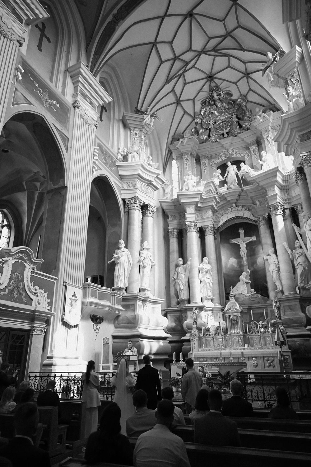 Black and white photo of a Catholic church interior during a wedding ceremony with ornate baroque altar and vaulted ceiling.