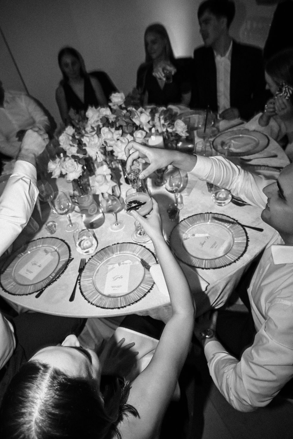 Black and white photo of elegantly dressed guests toasting at a formal dinner table with floral centerpiece.