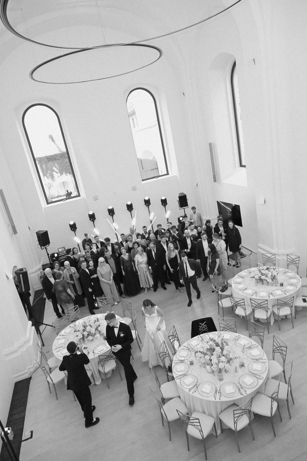 Aerial black and white photo of an elegant wedding reception in a white hall with round tables and gathered guests.