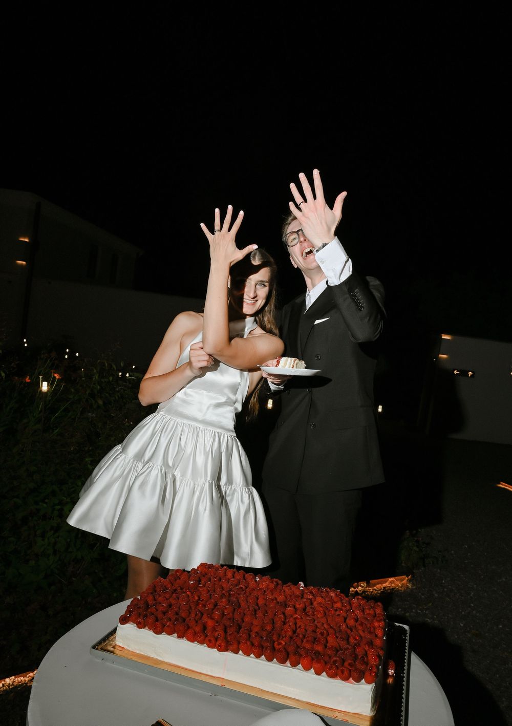 Bride and groom raising hands playfully beside a strawberry-topped wedding cake outdoors at night.
