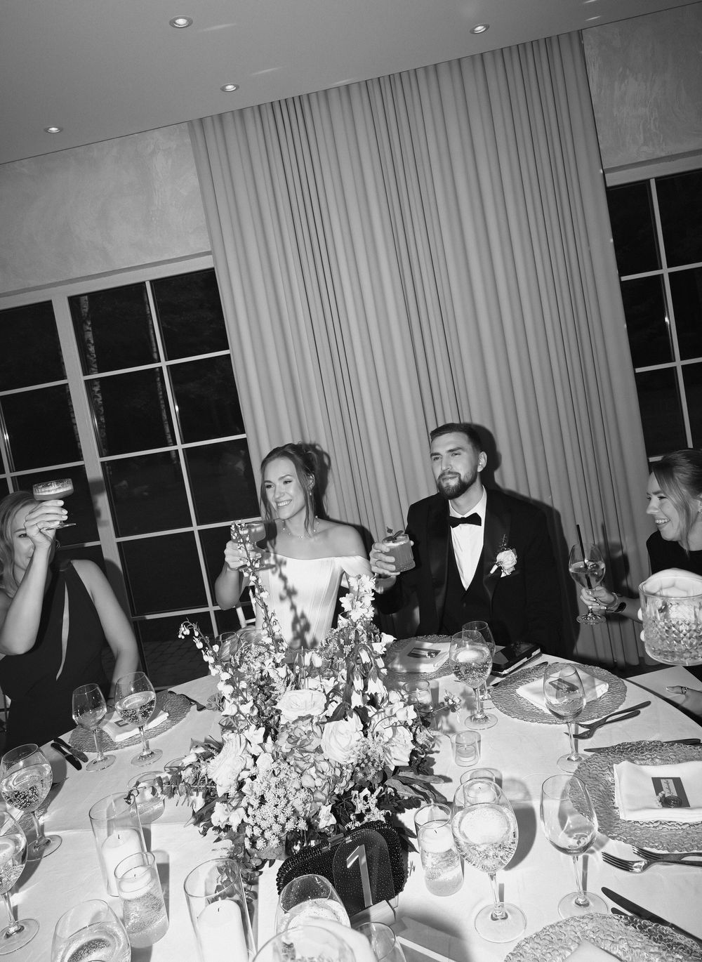Black and white wedding reception photo of bride and groom toasting at elegantly decorated table with floral centerpiece.