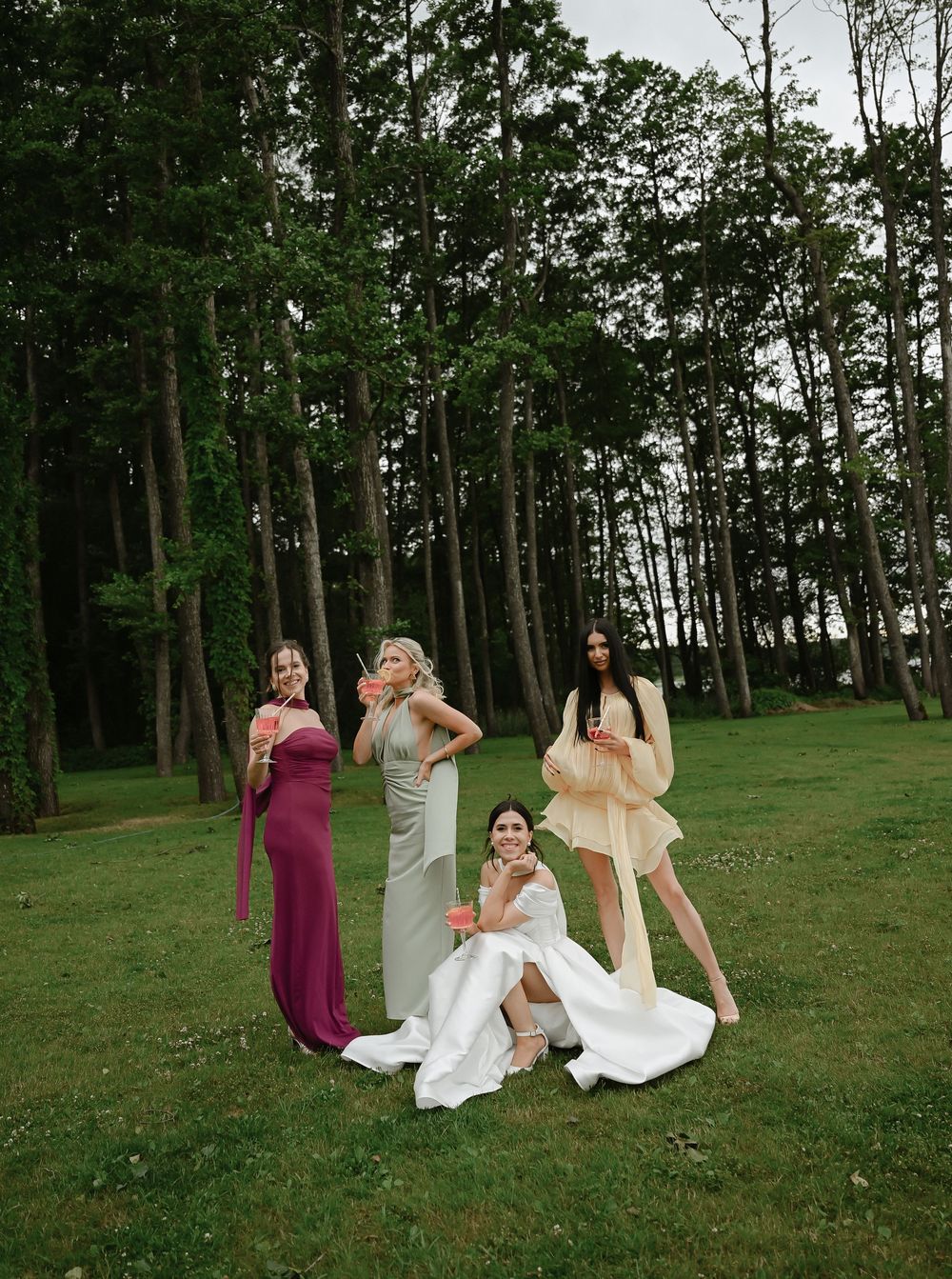 Bride in white gown poses with three bridesmaids in colorful dresses on lush green lawn surrounded by tall trees.