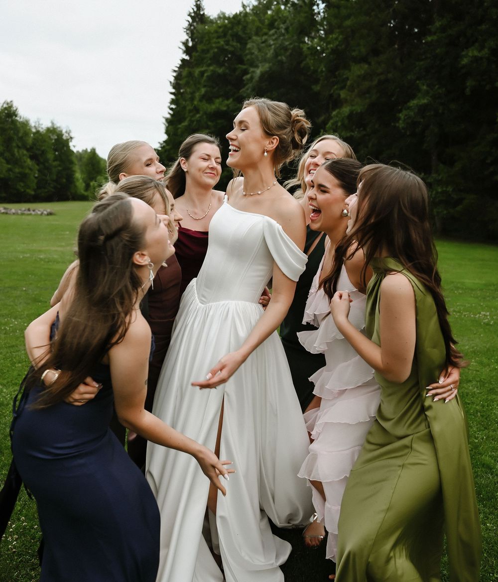 A bride in a white gown laughs joyfully surrounded by bridesmaids in colorful dresses on a green lawn.