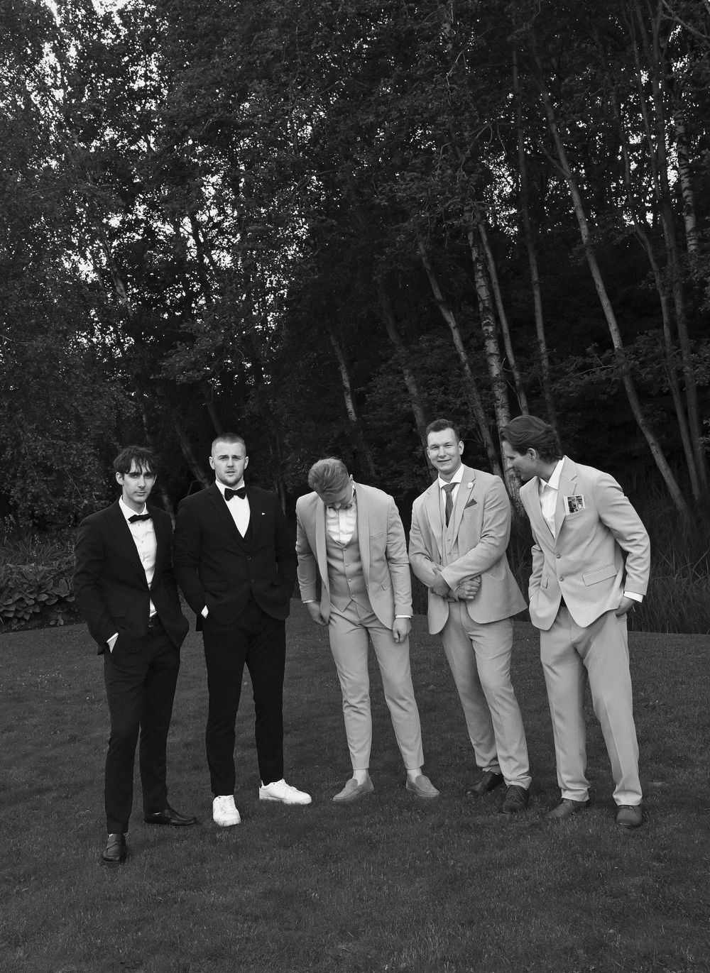 Five men in formal suits pose on a green lawn with tall trees in the background in this black and white photo.