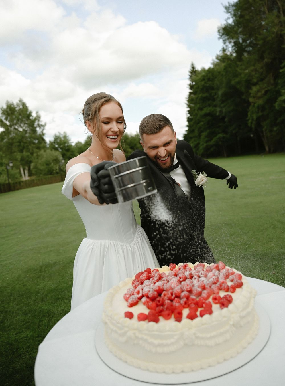 Bride and groom laughing while dusting powdered sugar over a white wedding cake topped with fresh raspberries outdoors.