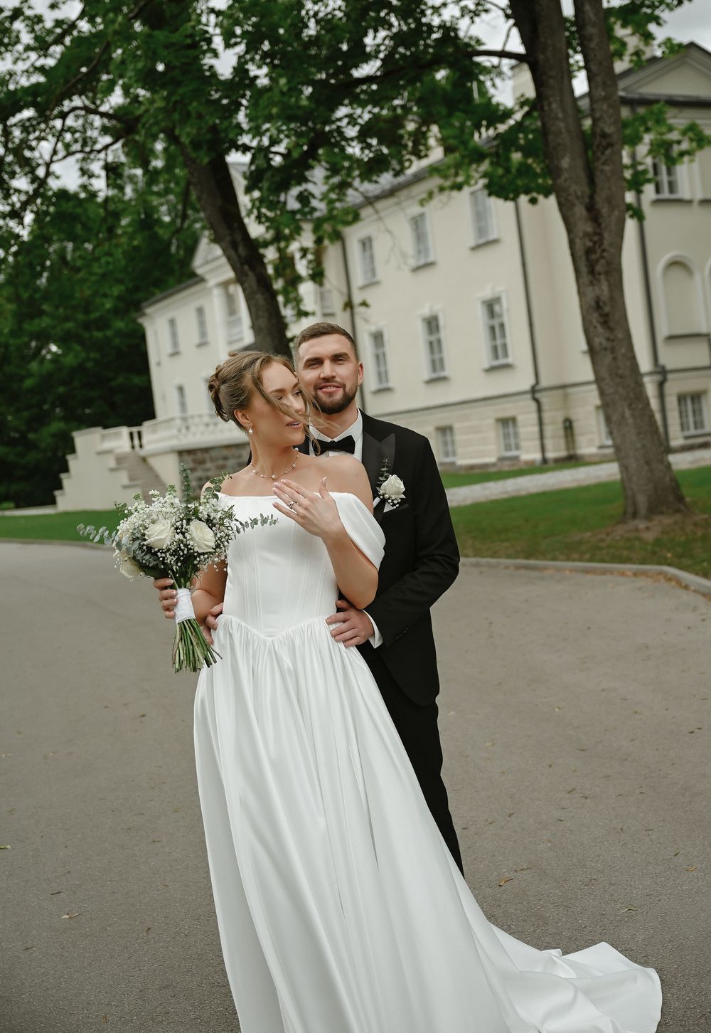 A bride in a white off-shoulder gown holds a bouquet as a groom in a black tuxedo embraces her outdoors.