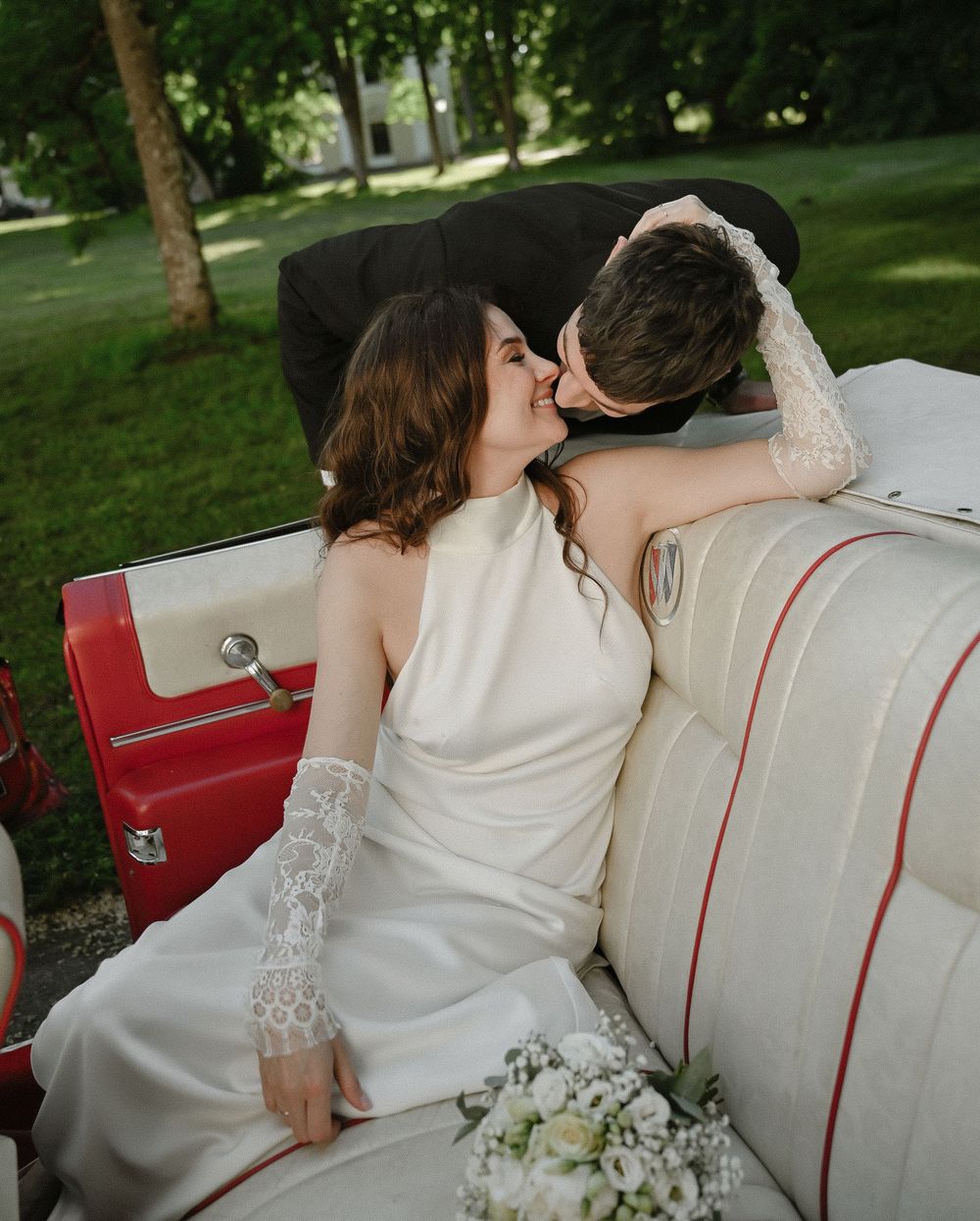 Bride and groom sharing a romantic kiss in a vintage red and white convertible car on their wedding day.