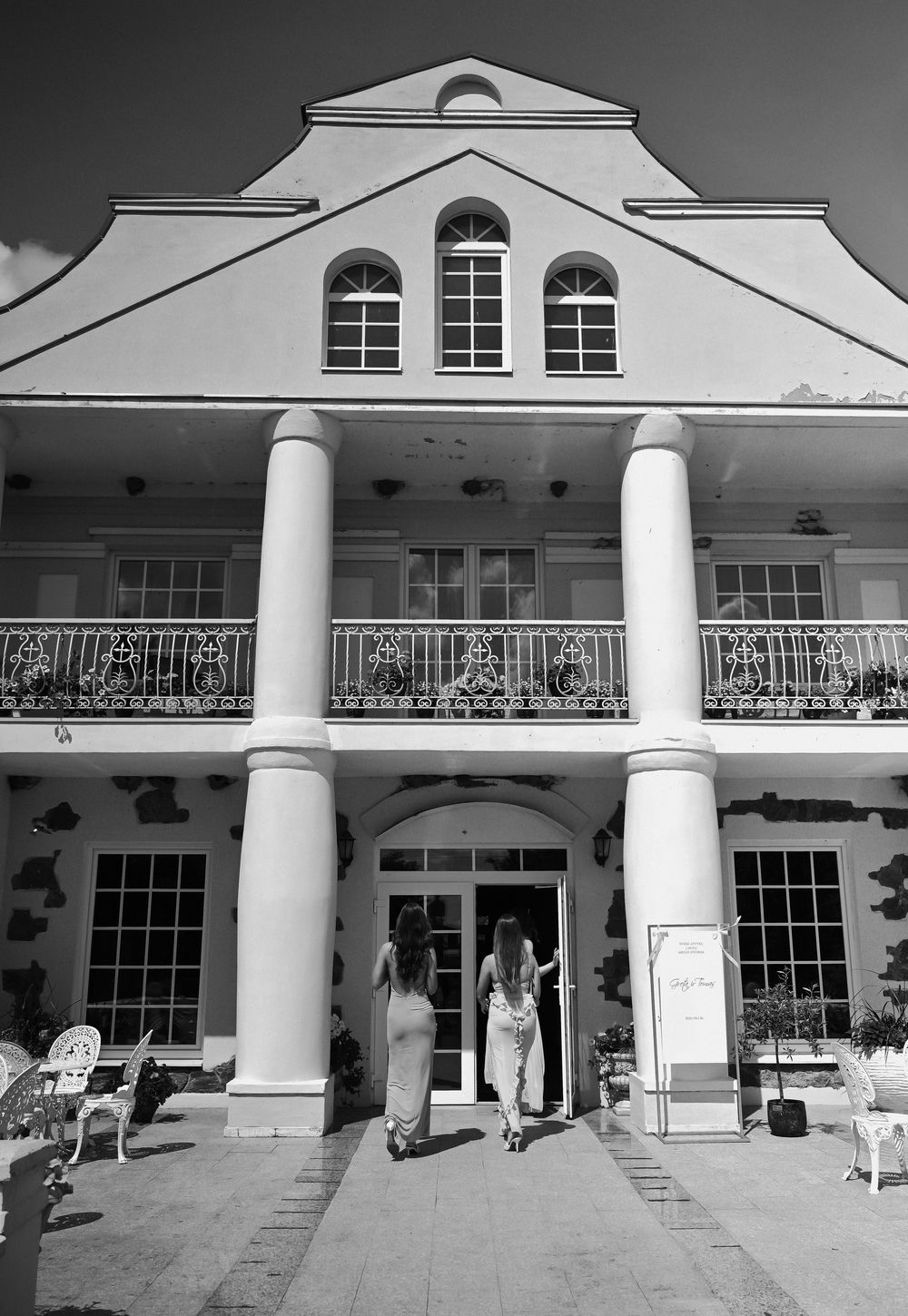 Black and white photo of two women entering an elegant colonial-style building with white columns and ornate balcony.
