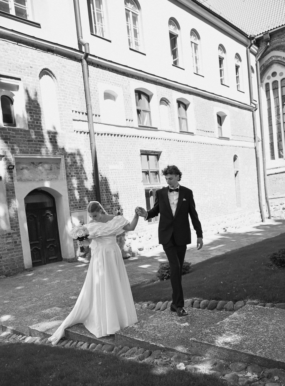 Black and white photo of a bride and groom holding hands walking outside a historic brick building on their wedding day.