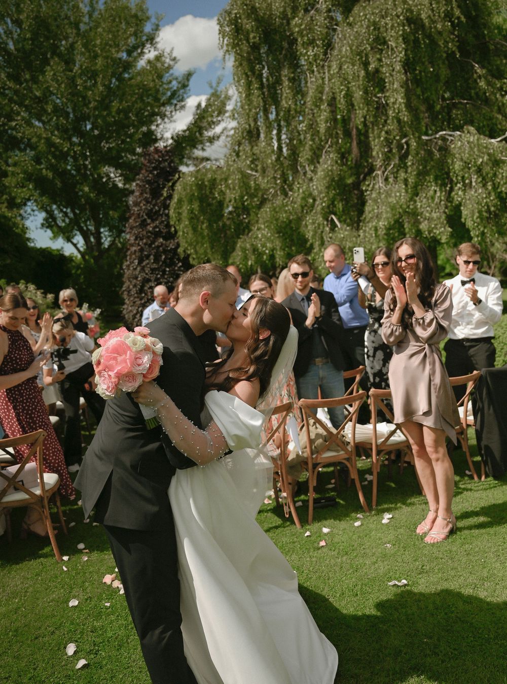 Newlywed couple sharing a kiss at outdoor wedding ceremony while guests celebrate on sunny garden lawn.