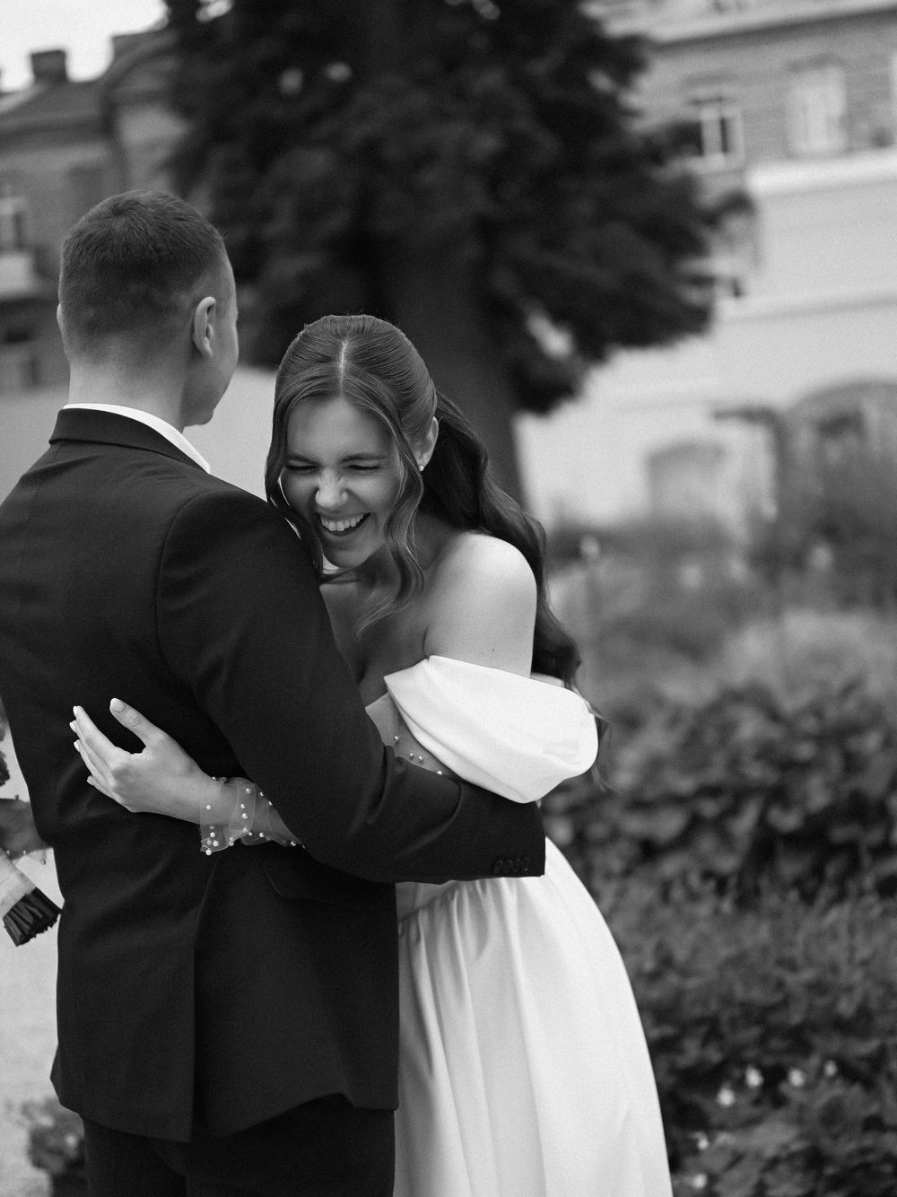 Black and white photo of a laughing bride in off-shoulder gown embracing groom in suit at outdoor wedding venue.