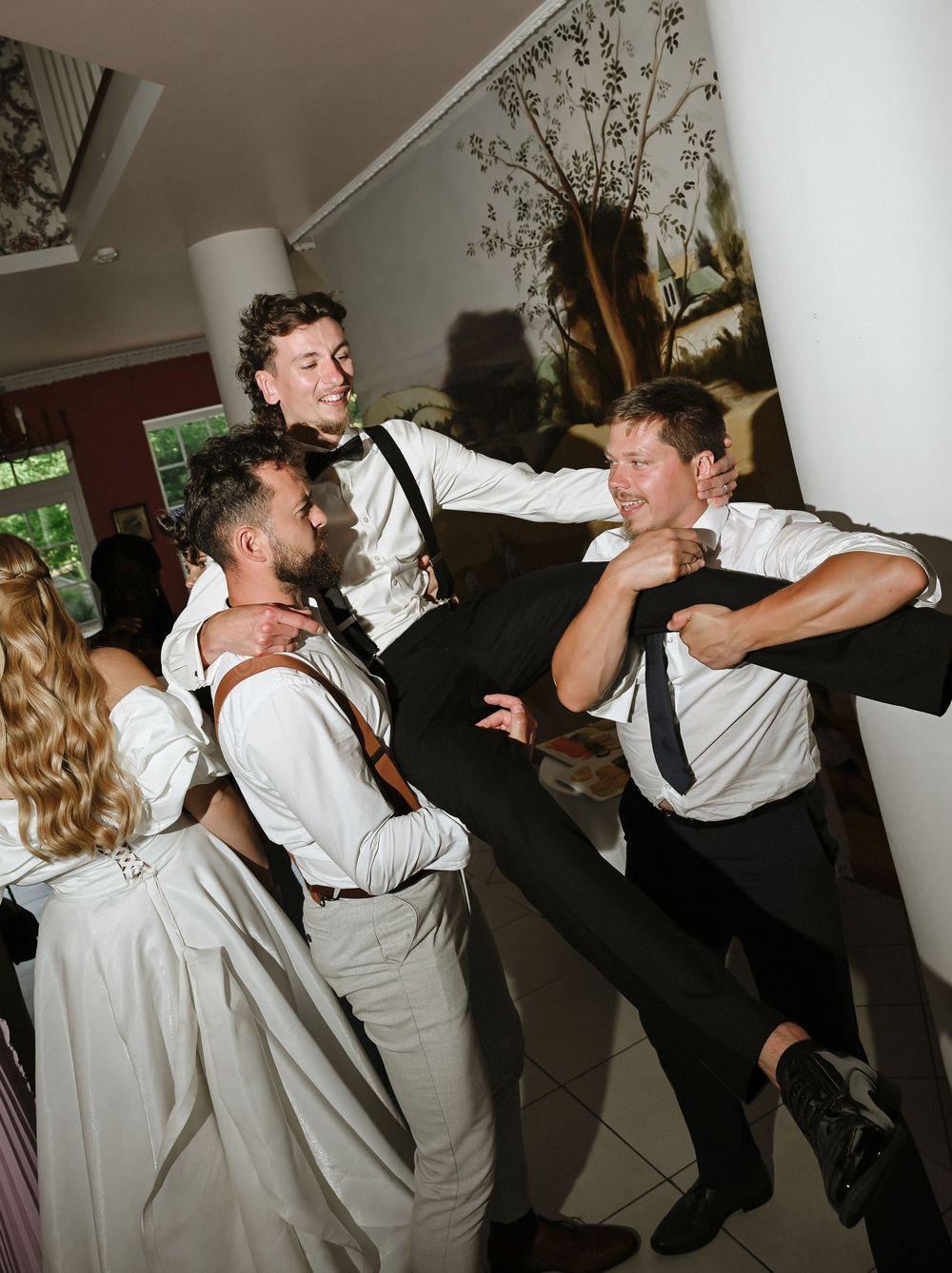 Groomsmen in white shirts and suspenders laughing and joking around at a wedding reception venue.