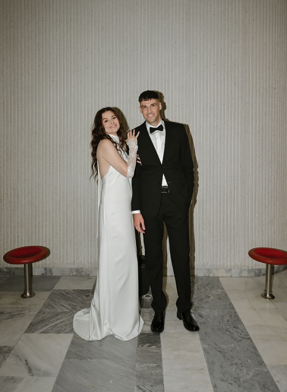 Couple posing in formal attire, bride in white gown and groom in black tuxedo, standing on marble floor.