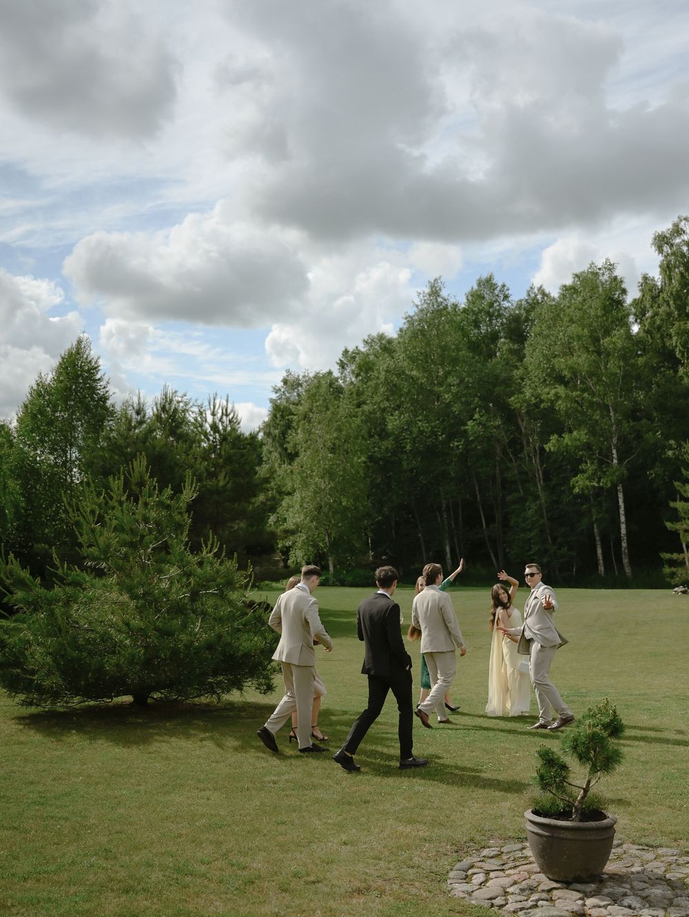 A wedding party walks across a lush green lawn surrounded by tall trees under a cloudy sky.