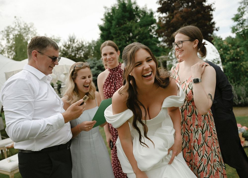 Bride in white dress laughing with wedding guests outdoors at a garden celebration.