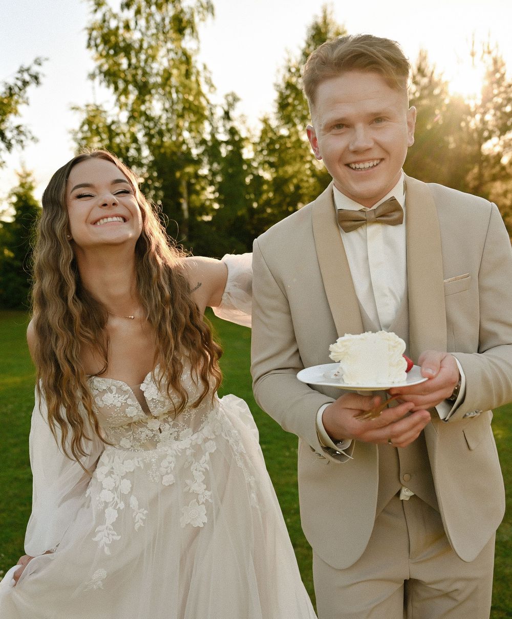Smiling bride in white lace gown and groom in tan suit holding wedding cake outdoors in sunlit garden.
