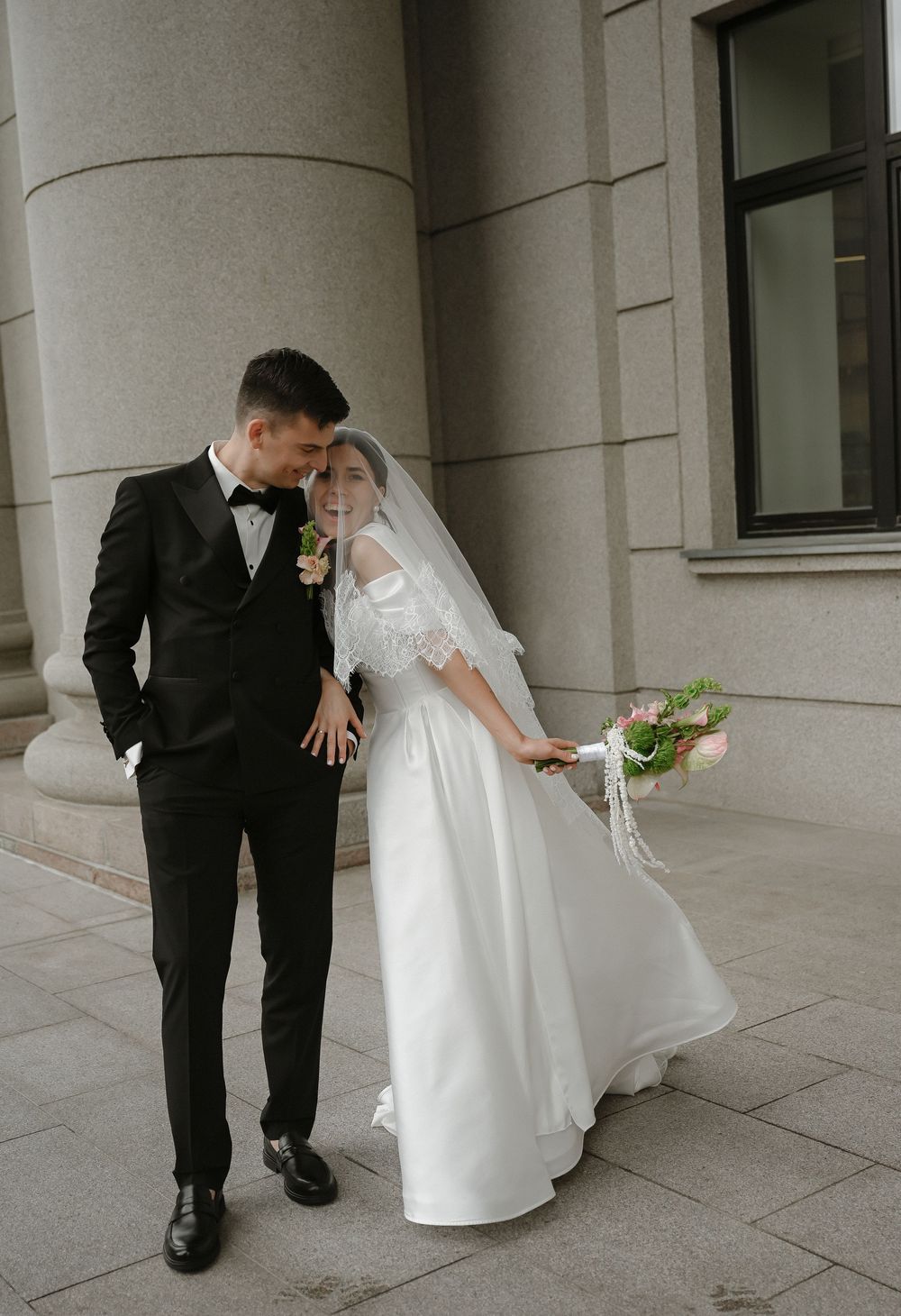 A couple in wedding attire poses by stone columns, groom in black tuxedo, bride in white gown holding a floral bouquet.