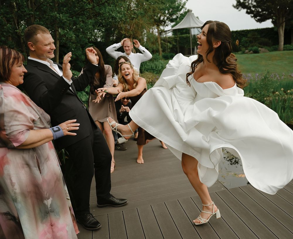 Bride in white gown twirling on deck surrounded by cheering wedding guests outdoors.