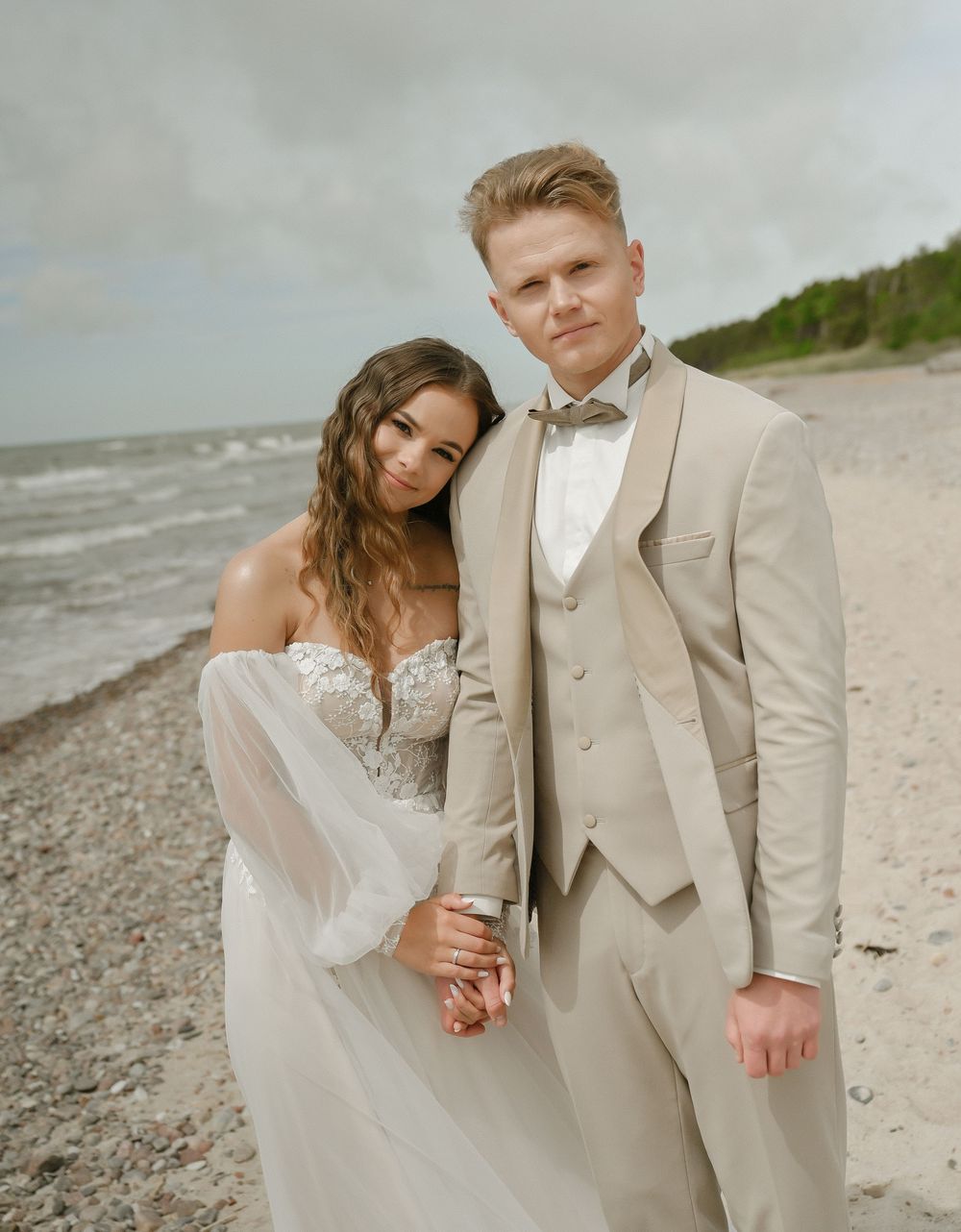 Couple in wedding attire posing on a beach, groom in beige suit and bride in white lace gown, ocean backdrop.