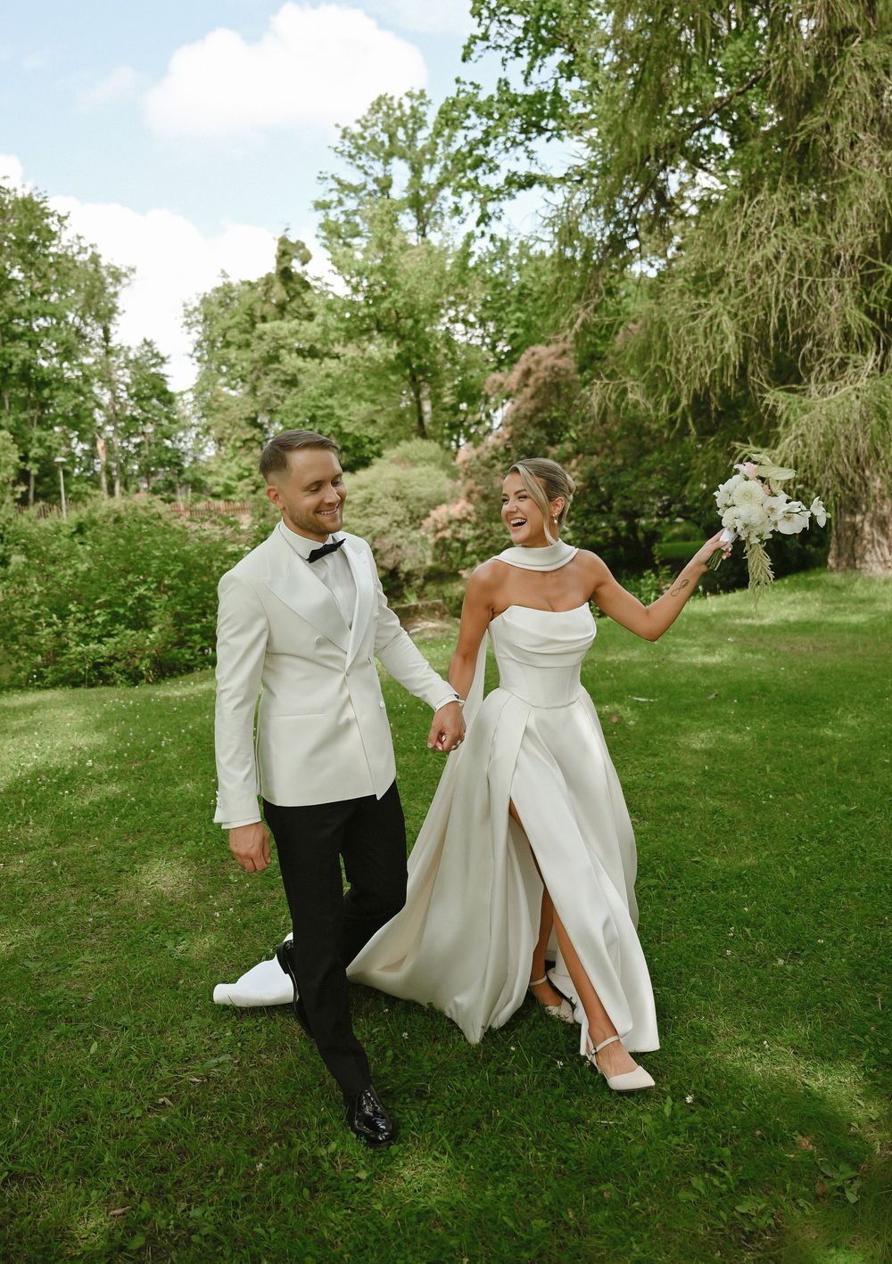 A bride and groom walk hand-in-hand through a lush green garden, smiling joyfully on their wedding day.