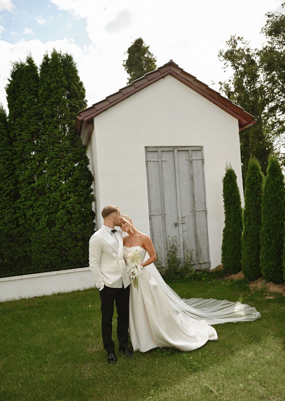 Bride and groom share a romantic kiss in front of a white cottage surrounded by tall evergreen trees on their wedding day.