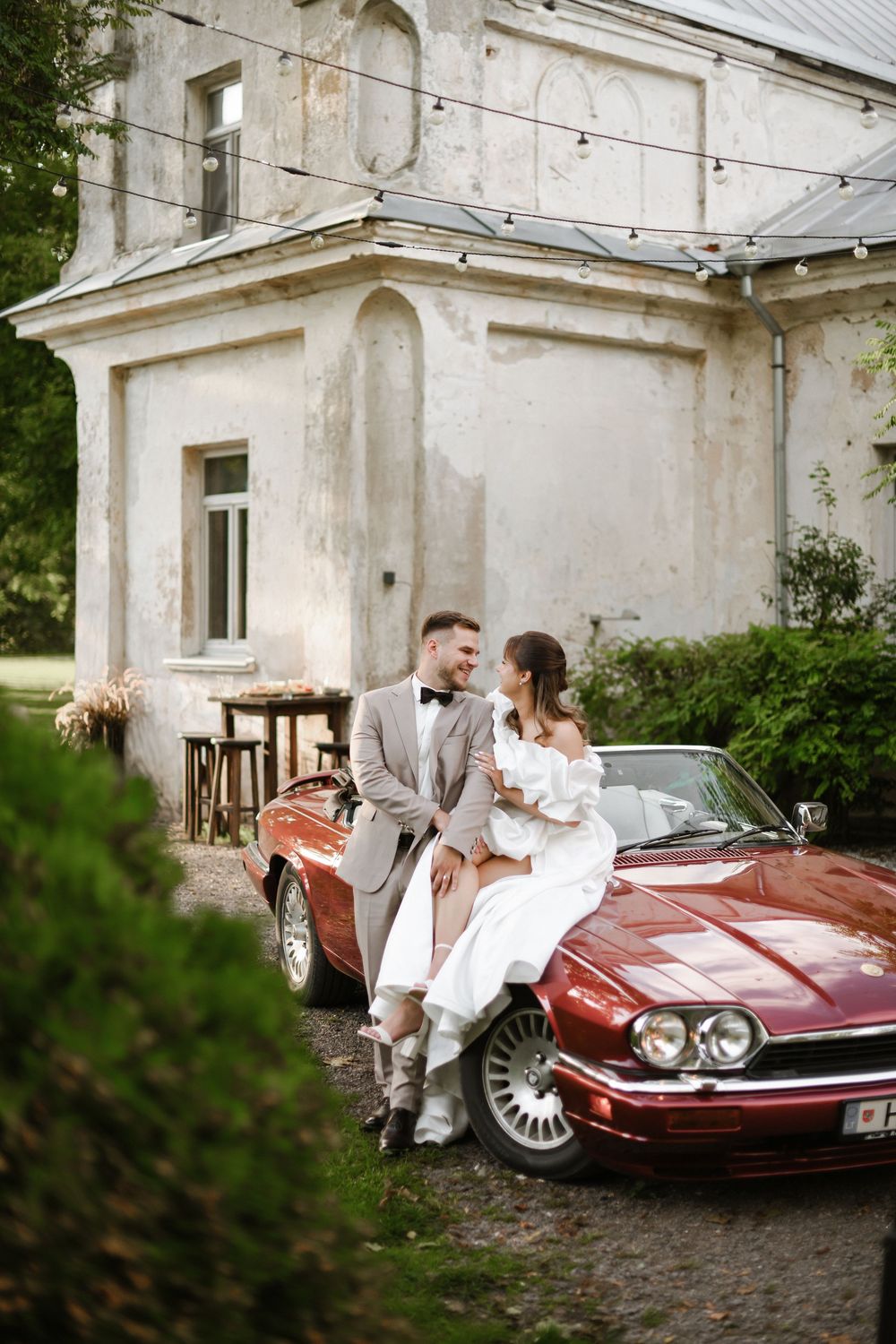 A couple poses romantically near a vintage burgundy car outside a rustic white building.