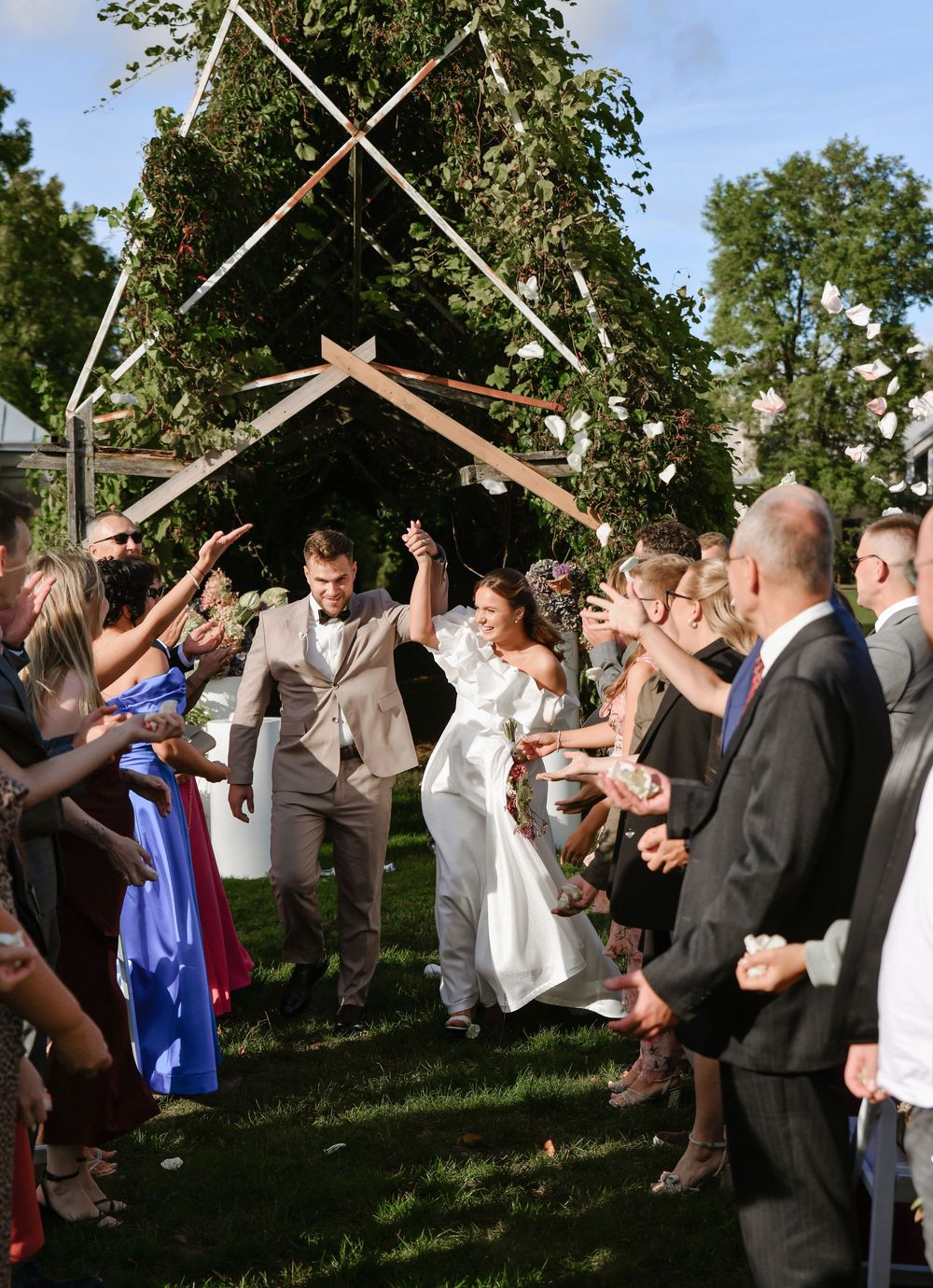 Wedding guests form a celebratory arch with raised arms as couple exits their outdoor garden ceremony.