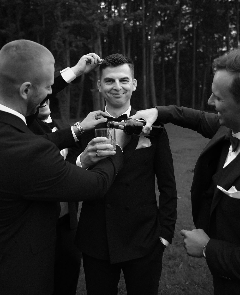 Black and white photo of groom being helped by groomsmen adjusting his bow tie and boutonniere before wedding.