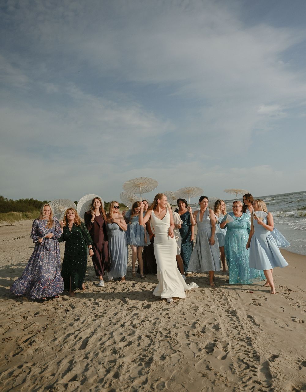 Bride in white gown poses with bridesmaids in colorful dresses on sandy beach, holding parasols under cloudy sky.