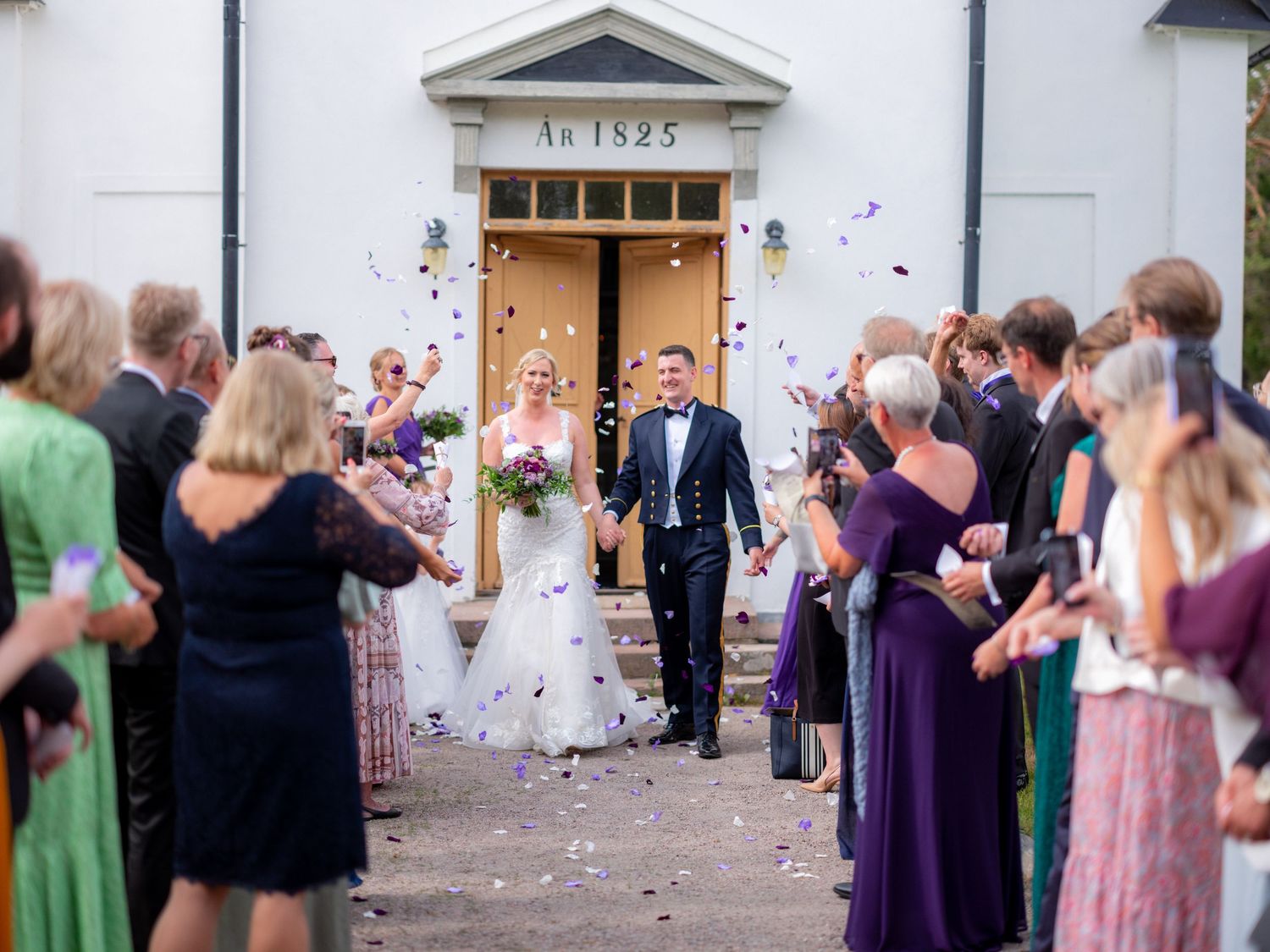 Wedding guests throwing petals as newlyweds exit white church building.