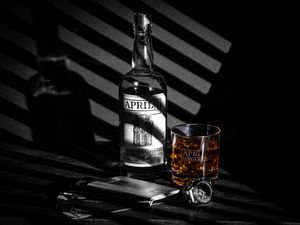 A bottle of Rebel bourbon whiskey stands next to a glass with ice in dramatic lighting with shadows from blinds.