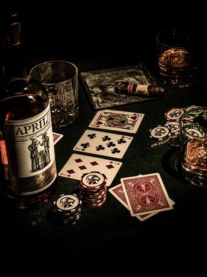 Playing cards, poker chips, and a bottle of Abril liquor arranged on a dark table in moody lighting for gambling.