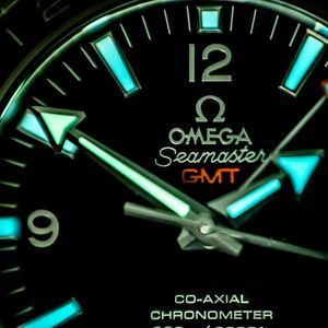 Close-up view of an illuminated Omega Seamaster GMT watch face showing blue-green lume and co-axial chronometer text.