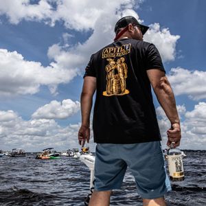 Person wearing black t-shirt with gold graphic standing on boat deck against blue sky. Editorial product photography. April One Bourbon