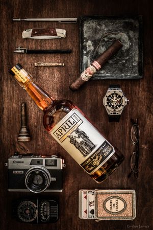 Flatlay of vintage items including bourbon, camera, watch, cigar and accessories on wooden surface.  Product photography. April One