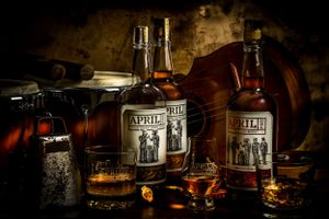 A rustic still life with whiskey bottles, glasses and cigar on dark wooden table with dramatic lighting.