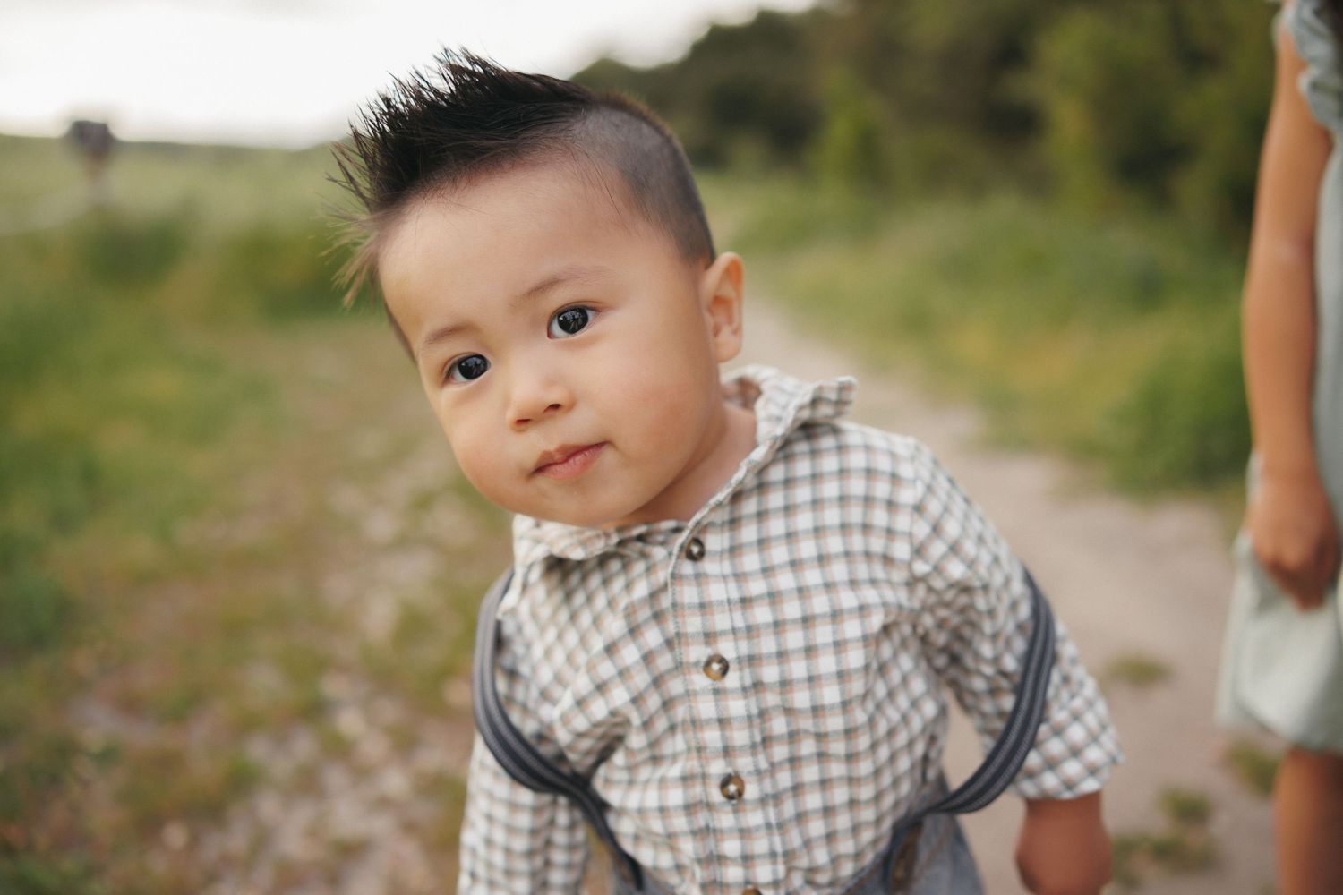 Adorable Asian toddler boy with a mohawk hairstyle wearing a plaid shirt, posing outdoors on a dirt path.