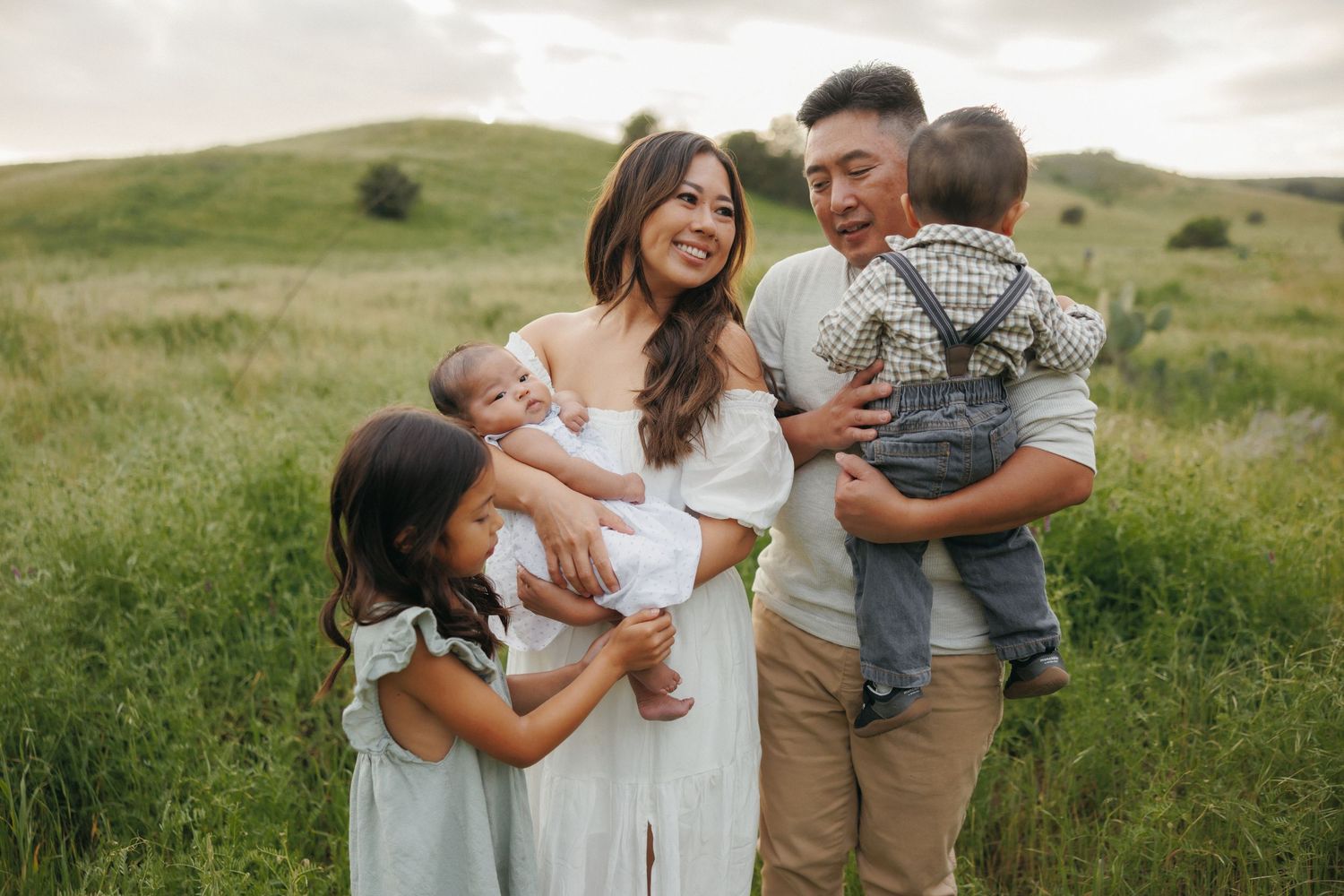 A happy Asian family of five poses outdoors in a lush green field, with parents holding young children.