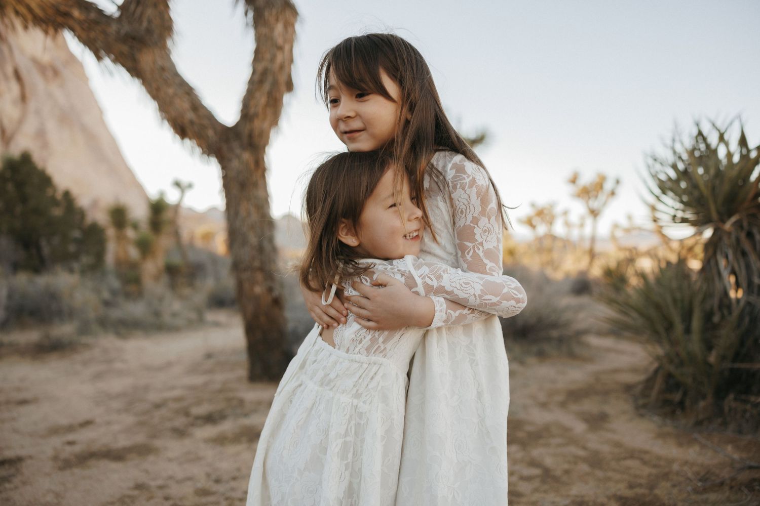 Two young girls in white lace dresses share a warm hug outdoors near Joshua trees in a desert landscape.