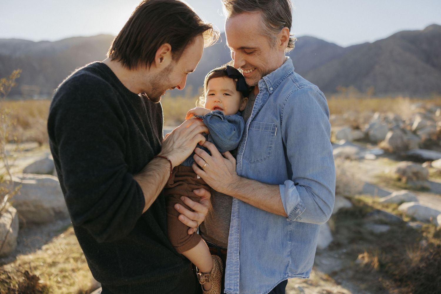 Two dads holding their baby outdoors in a rocky field with mountains in the background during golden hour.