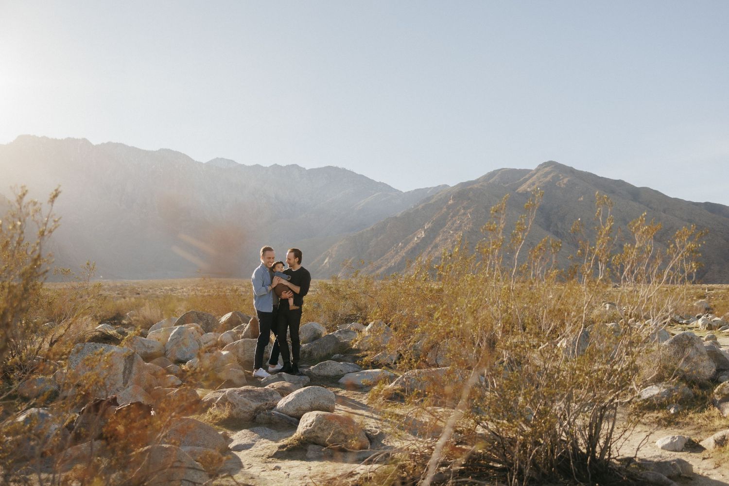 Couple embracing on rocks in a desert landscape with mountains and golden sunlight in the background.