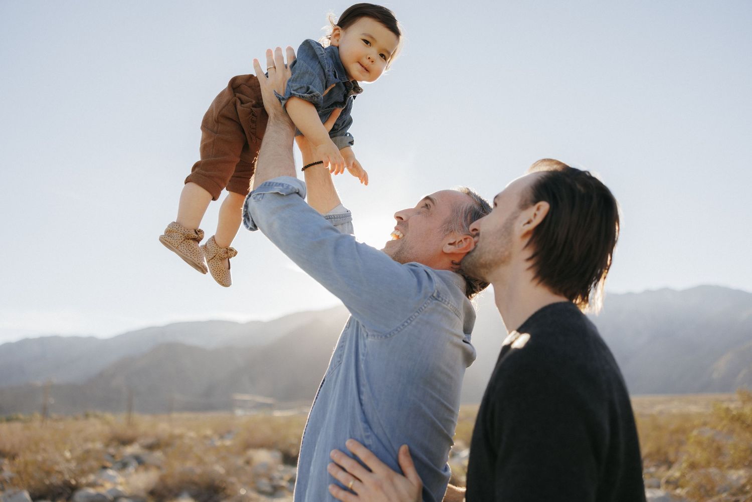 Two dads joyfully lifting their toddler in the air outdoors with mountains in the background on a sunny day.