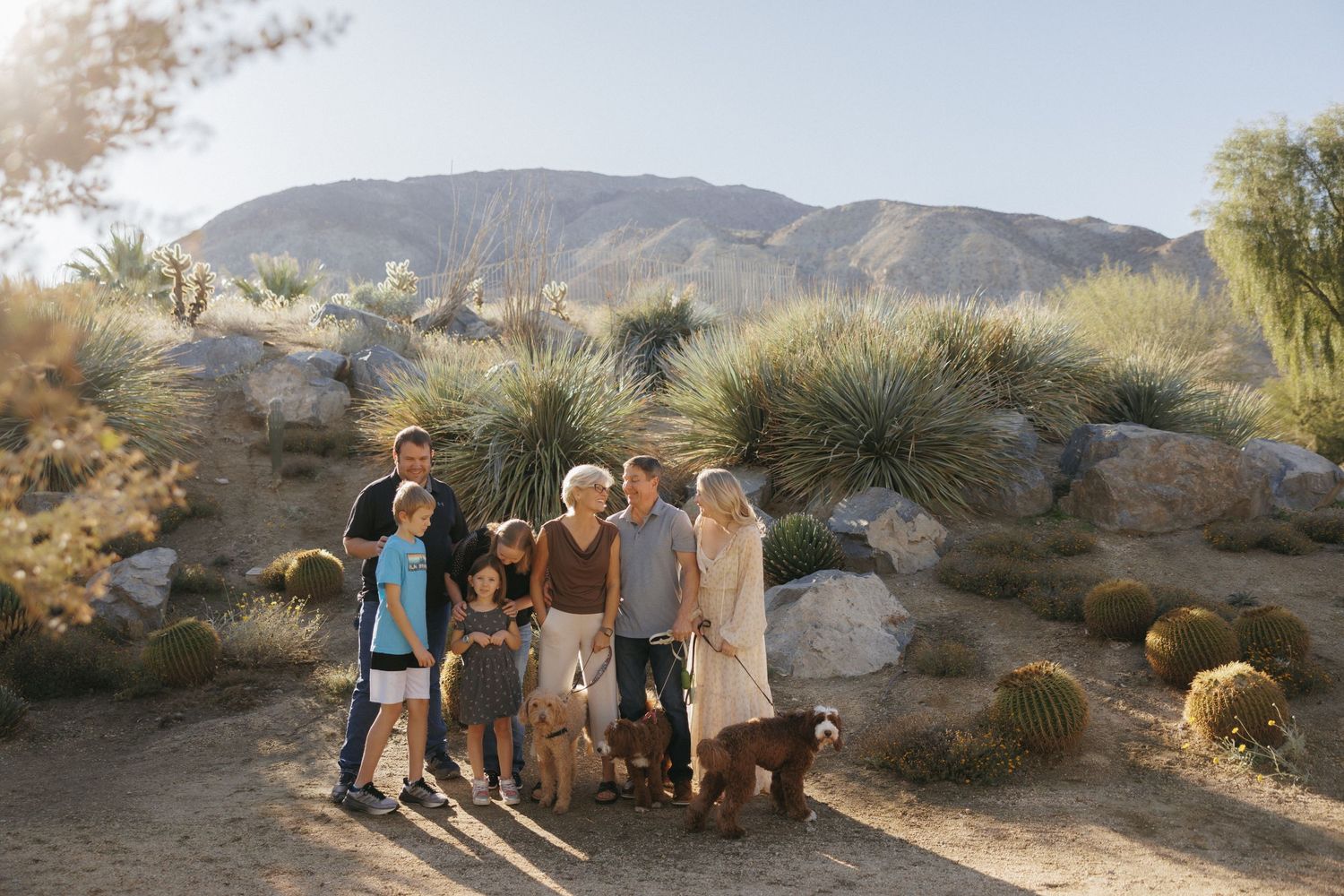 Family group portrait in desert landscape with mountains, cacti, and yucca plants in warm natural sunlight.
