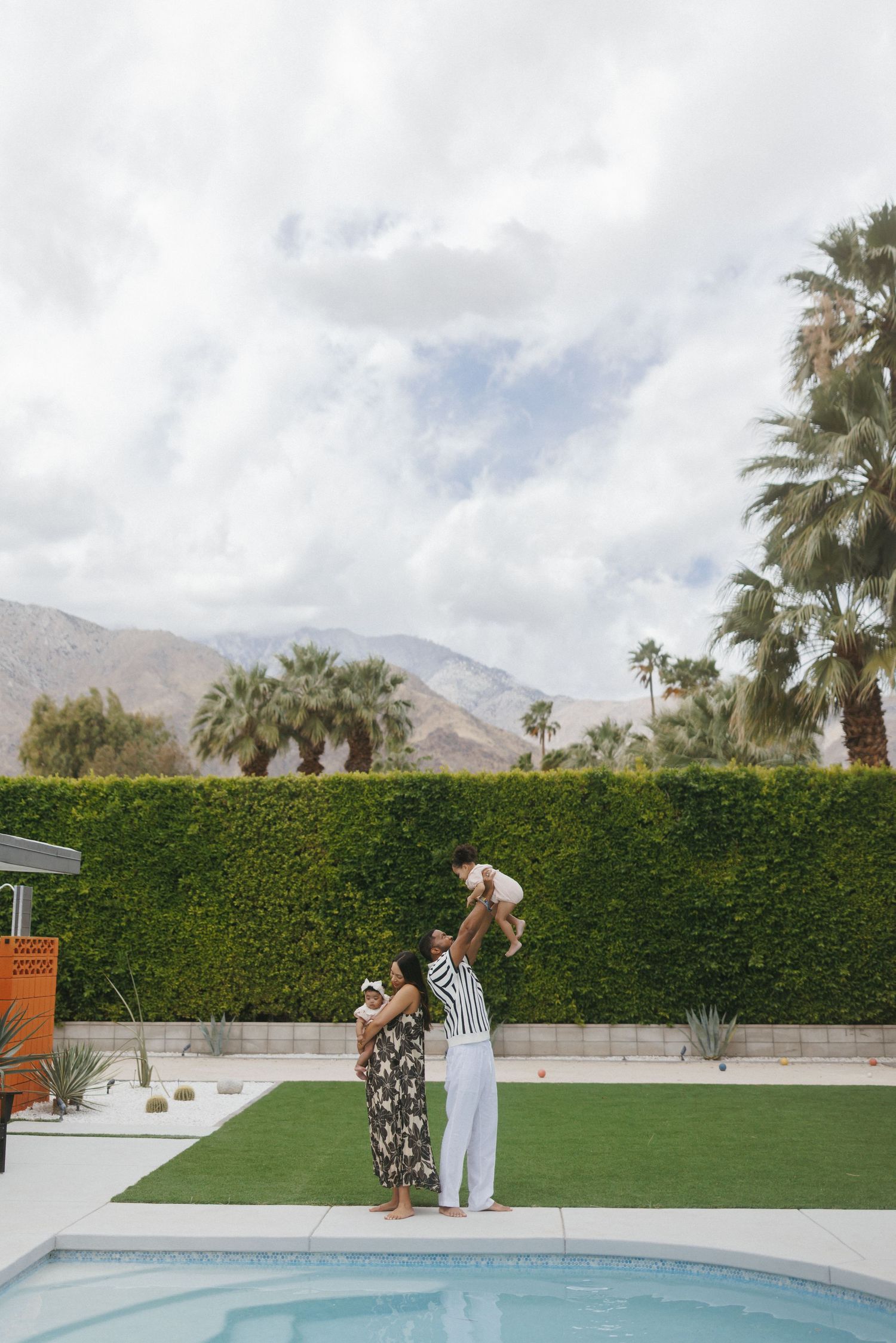 Family enjoying poolside time in Palm Springs, lifting baby in the air with palm trees and mountains in the background.