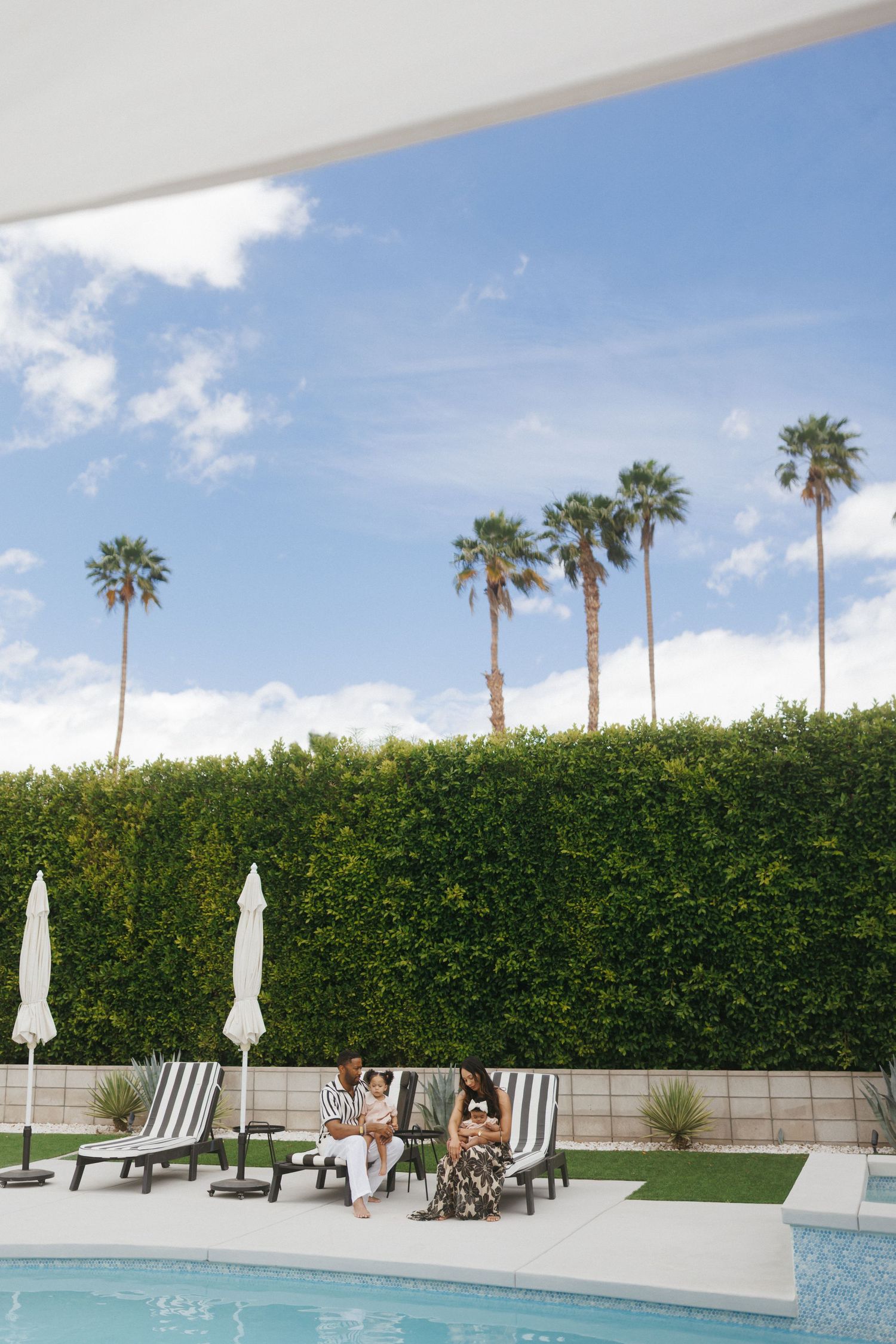 Two women relaxing on lounge chairs by a pool surrounded by tall hedges and palm trees under a blue sky.