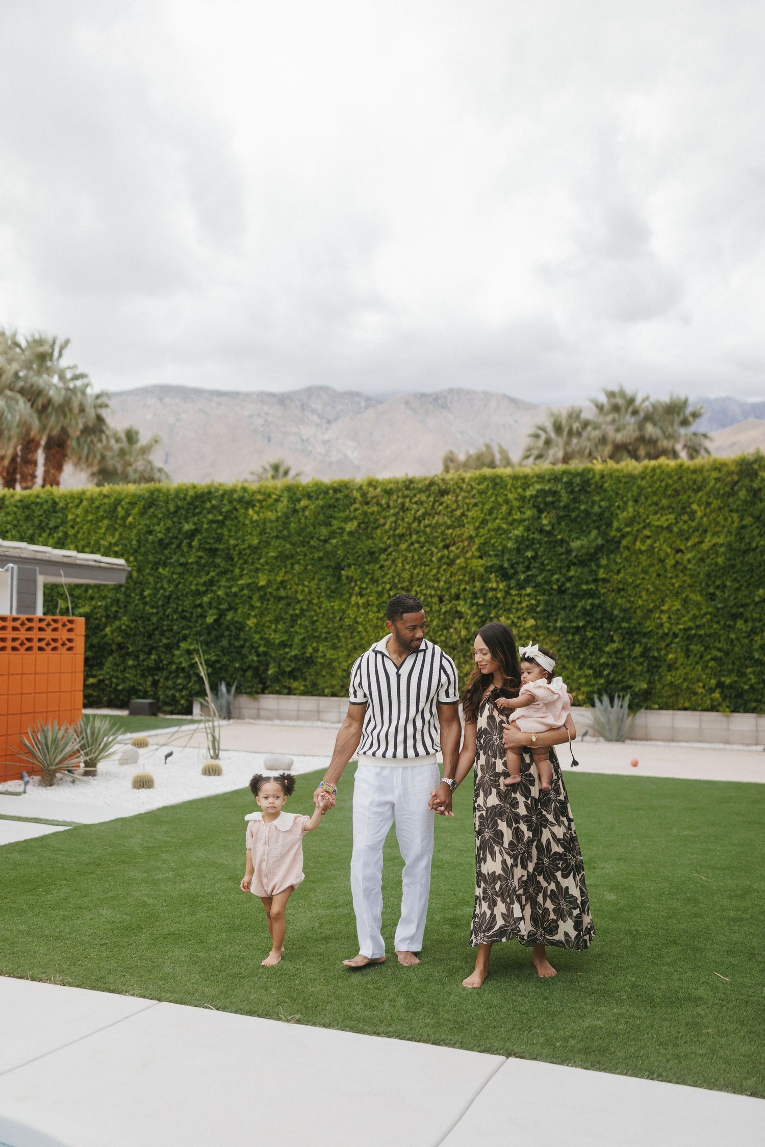 Family of four posing in lush backyard with mountain views, parents holding children on green lawn in Palm Springs.