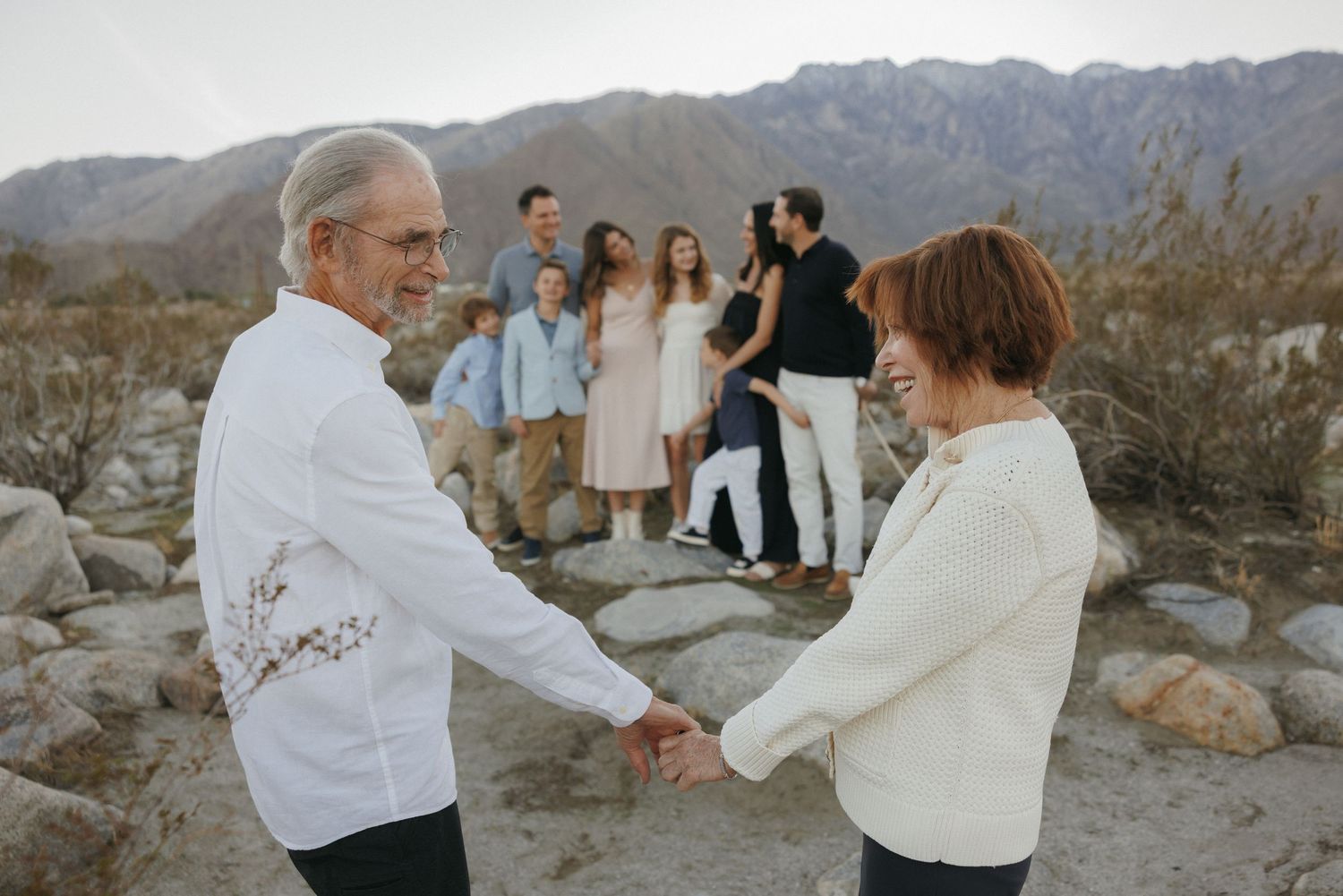 Elderly couple holds hands and dances outdoors with family gathered among desert rocks and mountains in background.