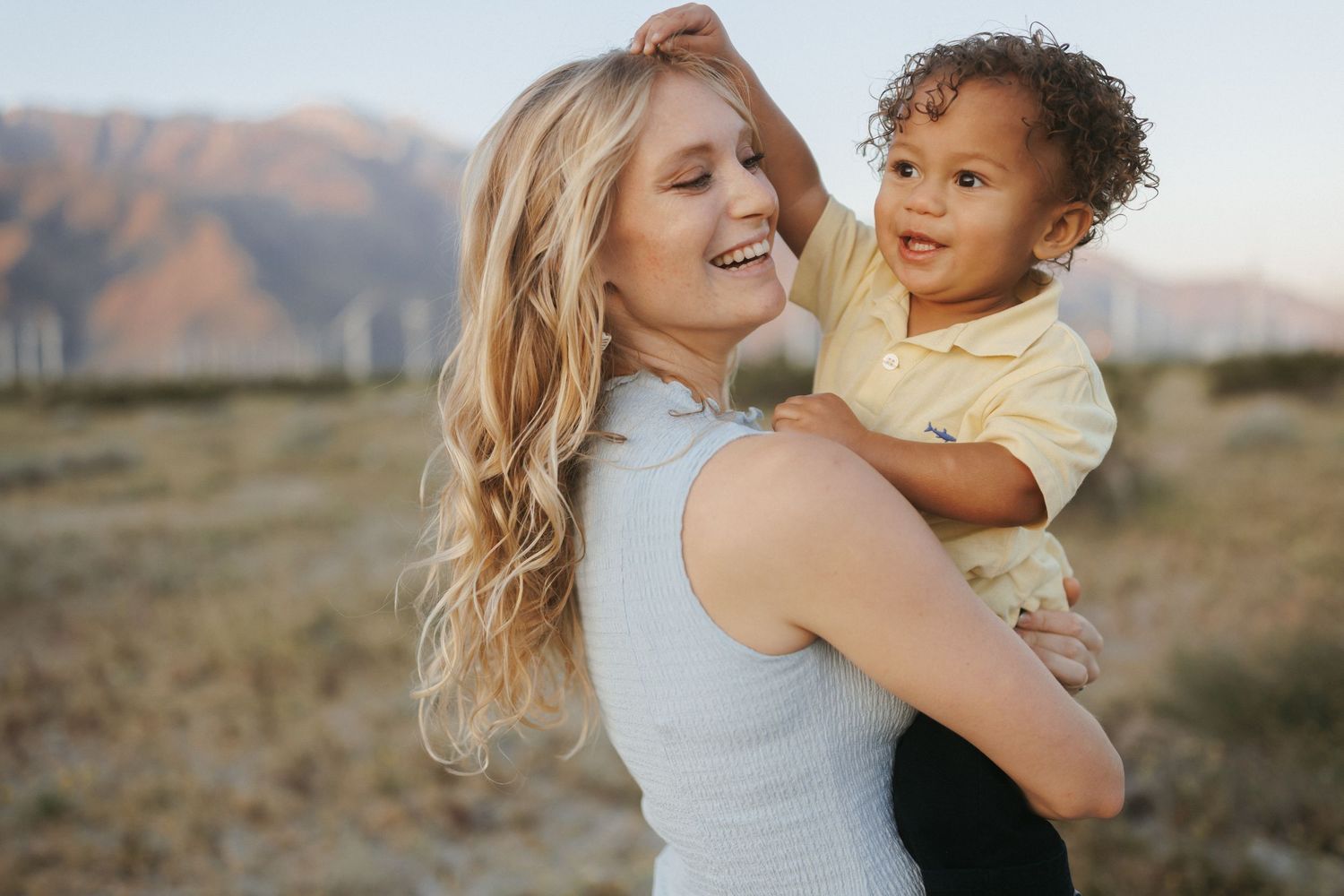Smiling blonde woman holding a curly-haired toddler outdoors with mountains in the background.