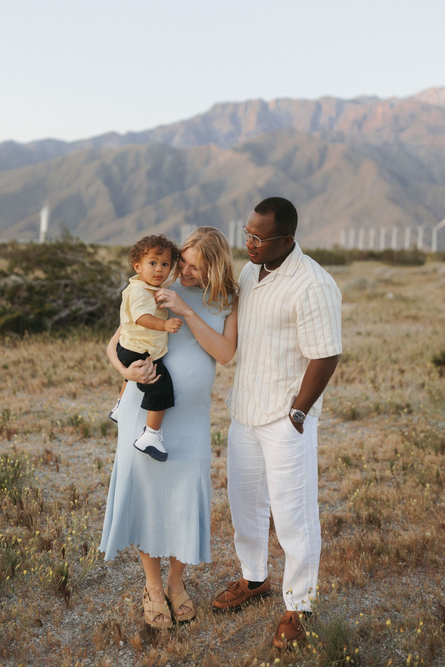 Interracial family posing outdoors in desert landscape with mountains and wind turbines in background.