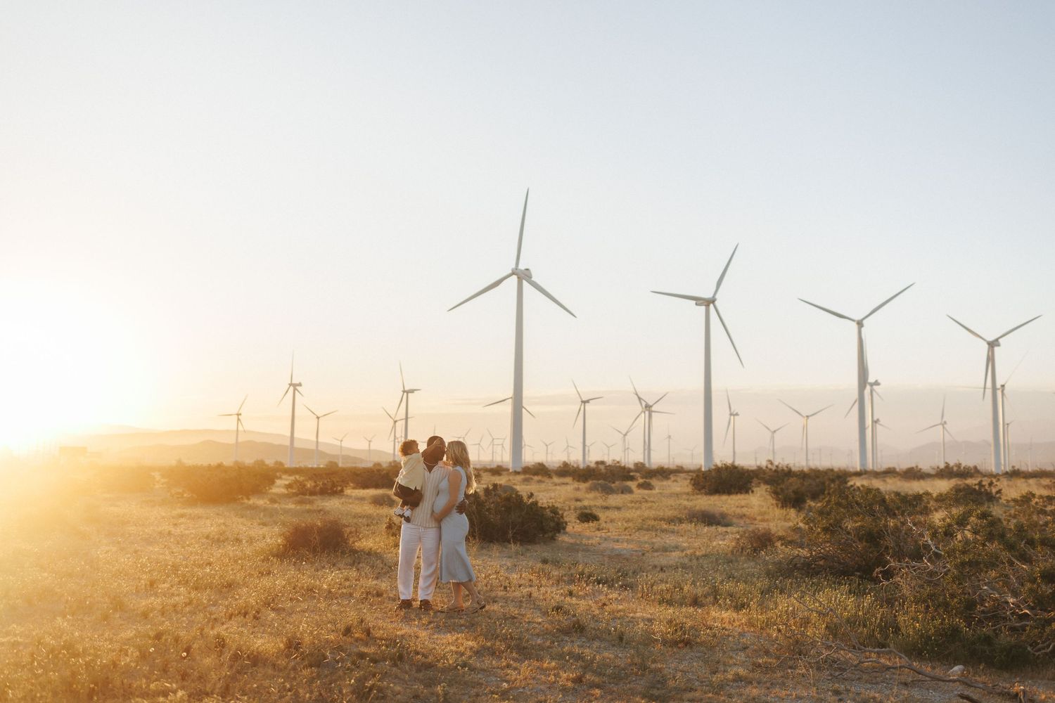 Couple embracing at sunset in a wind farm field with tall turbines against a golden sky.